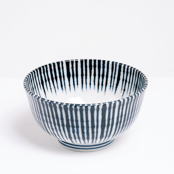 A side view of the white Mino-yaki Tokusa Noodle Bowl on show at NiMi Projects UK. The bowl is made of Japanese porcelain and featuring a traditional pattern of blue stripes representing horsetail grass that extends from its sides over the lip of the bowl to end on the inside in a zigzag pattern.