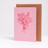A pink greeting card, with its brown envelope displayed behind, designed by Takako Copeland and available at NiMi Projects, featuring a letter-pressed printed illustration of a bouquet of flowers in red.