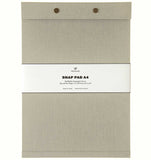 A warm grey fabric-covered A4 Postalco Snap Pad, showing the two roundmetal snap clasps at its top and a white label wrapped around its center featuring the Postalco bird logo.