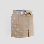 A short beige Sanpu Sanyo apron, as worn, with graphic patterned tenugui cotton pocketed side panels, a sailcloth canvas double-pocketed front and sanada himo woven waist cord. Made in Japan and available at NiMi Projects UK