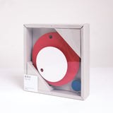 A Fukunaga Paper Clock of two circles, a small white one, on top of a red one, packed in a square bod with an additional small blue decorative dot, on display at NiMi Projects UK.