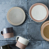 A collection of Angle mino-yaki earthenware products on diplay on a paved stone background. Featured are three kinds of Soil Plage, each with a central circle of rustic coloured glaze (from left to right - grey, white and taupe), and two Soil Mugs with tops dipped in glaze, one is graphite black and one in white. Image courtesy of Angle.