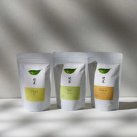 Three pouches of Awaido Japanese green tea leaves. The pouches are white and each have a green illustration of a tea leaf and the name of the tea in Japanese kanji. Below the kanji are coloured rectangle labels (yellow, green and orange), each with the name of the tea in English — Sumi, Iro and Kuan. Image ©AWAIDO