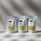 Three pouches of Awaido Japanese green tea leaves. The pouches are white and each have a green illustration of a tea leaf and the name of the tea in Japanese kanji. Below the kanji are coloured rectangle labels (yellow, green and orange), each with the name of the tea in English — Sumi, Iro and Kuan. Image ©AWAIDO