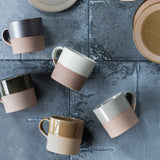 A collection of five mino-ware mugs, made in Japan. Each mug's top half is dipped in a colored glaze, clockwise from the top left — black, white, gray, brown and deep green.