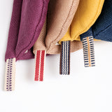 A closeup detail image of the bottoms of four hanging Ban Inoue pot holders (oven mitts) in different colours. Each holder is semicircle in shape and features a braided hanging cord at the bottom. Colours from left to right: burgundy, brown, yellow and navy blue. Made in japan and on show at NiMi Projects UK.