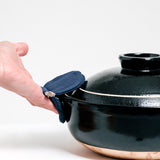 A navy blue Ban Inoue pot holder oven mitt being used to hold the side of a large brown Japanese nabe bowl. The pot holder is an oval shape, folded and stitched in the center to feature two semi-circle sides, is made from a lightly padded muslin and features a braided cord for hanging. Image by NiMi Projects UK.