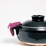A burgundy Ban Inoue pot holder oven mitt being used to hold the side of a large brown Japanese nabe bowl. The pot holder is an oval shape, folded and stitched in the center to feature two semi-circle sides, is made from a lightly padded muslin and features a braided cord for hanging. Image by NiMi Projects UK.