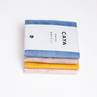 Four folded and stacked Caya muslin dish cloths or kitchen cloths, made in Japan by Ban Inoue using traditional mosquito-net weaving techniques. Each one is wrapped with a white, paper label slip and a different colour - from top to bottom: pale blue, beige, yellow and pastel pink. Available at NiMi Projects UK.