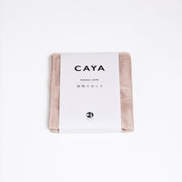 A folded beige Caya muslin dish cloth or kitchen cloth, made using Japanese cotton mosquito net weaving techniques by Ban Inoue. The cloth is wrapped in a white, paper label featuring the Caya brand name and it’s available at NiMi Projects UK.