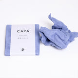 A folded pale blue Caya muslin dish cloth or kitchen cloth, made using Japanese cotton mosquito net weaving techniques by Ban Inoue. The cloth is wrapped in a white, paper label featuring the Caya brand name and is placed next to a washed and fluffed version. On show at NiMi Projects.