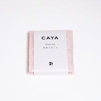 A folded pink Caya muslin dish cloth or kitchen cloth, made using Japanese cotton mosquito net weaving techniques by Ban Inoue. The cloth is wrapped in a white, paper label featuring the Caya brand name and it’s available at NiMi Projects UK.