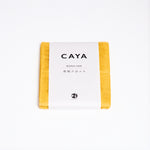 A folded yellow Caya muslin dish cloth or kitchen cloth, made using Japanese cotton mosquito net weaving techniques by Ban Inoue. The cloth is wrapped in a white, paper label featuring the Caya brand name and it’s available at NiMi Projects UK.
