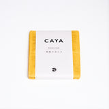 A folded yellow Caya muslin dish cloth or kitchen cloth, made using Japanese cotton mosquito net weaving techniques by Ban Inoue. The cloth is wrapped in a white, paper label featuring the Caya brand name and it’s available at NiMi Projects UK.