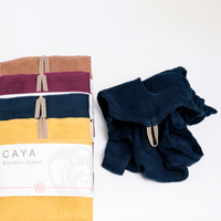Four different coloured, muslin Caya kitchen towels, fanned out vertically and placed alongside a navy blue washed and version. Each tea towel is made using traditional mosquito net weaving techniques and features a braided loop for hanging at the top. Each is wrapped in a white, paper slip with the Caya logo and is on show at NiMi Projects UK.