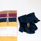 Four different coloured, muslin Caya kitchen towels, fanned out vertically and placed alongside a navy blue washed and version. Each tea towel is made using traditional mosquito net weaving techniques and features a braided loop for hanging at the top. Each is wrapped in a white, paper slip with the Caya logo and is on show at NiMi Projects UK.