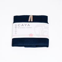 A folded navy blue Caya kitchen towel, made from Japanese muslin woven using traditional mosquito net weaving techniques. The tea towel is made in Japan by Ban Inoue and wrapped in a white paper label with the Caya logo. Available at NiMi Projects UK.
