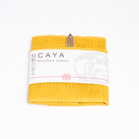 A folded yellow Caya kitchen towel, made from Japanese muslin woven using traditional mosquito net weaving techniques. The tea towel is made in Japan by Ban Inoue and wrapped in a white paper label with the Caya logo. Available at NiMi Projects UK.