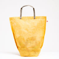 A large yellow muslin Vegetable tote bag, made in Japan by Ban Inoue using traditional mosquito net weaving techniques. The bag is filled with onions and has a folded and stitched base for support and short top handles. On show at NiMi Projects UK.