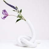 A large white Ceramic Japan Musubi Vase, designed by Makoto Komatsu, made from bisque porcelain shaped into a long tube tied in a loose knot, which acts as the base, and displaying purple flowers. On show at NiMi Projects UK.