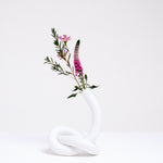 A white Ceramic Japan Musubi Vase, designed by Makoto Komatsu, made from bisque porcelain shaped into a long tube tied in a loose knot, which acts as the base, and displaying pink and purple flowers. On show at NiMi Projects UK.