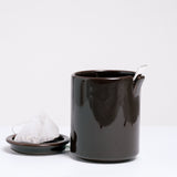 A brown Ceramic Japan Peel Cup, designed by Oki Sato of Nendo, featuring a small section that appears to be peeled away from the lip. A spoon rests in the peeled away section. On the left is a small dish holding a tea bag, which can also be used as a lid to the cup. Available at NiMi Projects UK.