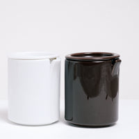 A pair of Ceramic Japan Peel Cups (left brown, right white), designed by Oki Sato of Nendo, each featuring a small section that appears to peeled away from the lip and indented lids. The section is to rest a spoon inside the cup. On display at NiMi Projects UK.