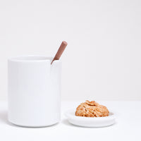 A white Ceramic Japan Peel Cup, designed by Oki Sato of Nendo, featuring a small section that appears to be peeled away from the lip. A spoon rests in the peeled away section. On the right is a small dish holding a biscuit, which can also be used as a lid to the cup. Available at NiMi Projects UK.