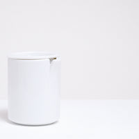 A white Ceramic Japan Peel Cup, designed by Oki Sato of Nendo, featuring a small section that appears to be peeled away from the lip. A lid, which can also be used as a small dish, keeps the beverage inside. Available at NiMi Projects UK.