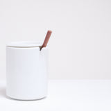 A white Ceramic Japan Peel Cup, designed by Oki Sato of Nendo, featuring a small section that appears to be peeled away from the lip. A spoon rests in the peeled away section. A lid, which can also be used as a small dish, keeps the beverage inside. Available at NiMi Projects UK.