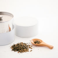 A Japanese hinoki (Japanese Cypress) wood tea  scoop beside a small pile of green tea leaves and a white Moheim tea caddy on display at NiMi Projects UK.