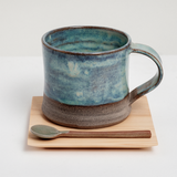 A handmade ceramic, earthenware mug with a blue-green drip glaze and strip-like handle and a matching ceramic tea spoon,  displayed on a large square Legnatec hinoki (Japanese cypress) wood coaster at 12cm x 12cm, with a tapered base. Available at NiMi Projects UK.