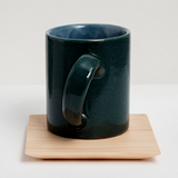 A deep green mug with an angled handle, displayed on a large square Legnatec hinoki (Japanese cypress) wood coaster at 12cm x 12cm, with a tapered base. On show at NiMi Projects UK.