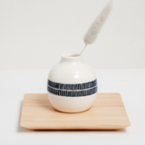 A white, small rotund bud vase with a navy blue vertically striped band around its centre, displayed on a large square Legnatec hinoki (Japanese cypress) wood coaster at 12cm x 12cm, with a tapered base. On show at NiMi Projects UK.