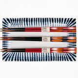 An aerial view of the rectangular NiMi Projects’ Mino-Yaki Tokusa Sushi Plate, with three pairs of wooden chopsticks in red and black on top. Made of white porcelain, the plate features a traditional Japanese horsetail grass pattern of indigo-blue zig-zag stripes that taper inward around the edge like a frame.