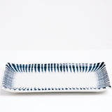 A side view of the rectangular Mino-Yaki Tokusa Sushi Plate, showing its upturned sides. Made of white porcelain, the plate features a traditional Japanese horsetail grass pattern of indigo-blue zig-zag stripes that taper inward around the edge like a frame. On show at NiMi Projects UK.
