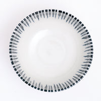 An aerial view of NiMi Projects’ Mino-Yaki Tokusa Noodle Bowl, a white Japanese porcelain piece, featuring a blue striped pattern of horsetail grass that extends over the rim to decorated the inside with a jagged, zigzag pattern.