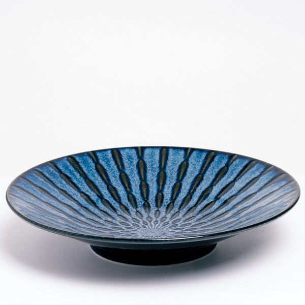 A side view of NiMi Projects’ Mino-Yaki Tokusa Blue Platter, featuring an indigo blue overglaze and a radial striped pattern of black underglaze, exposed by indents in the porcelain. The plate section stands on a 5cm tall base.