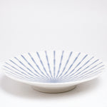 A side view of the NiMi Projects Mino-Yaki Tokusa White Platter, showing its 5cm-tall base. The plate features a pale blue striped pattern that radiates fro the center and is achieved by the pooling of blue glaze in subtle indentations of the ceramic. 