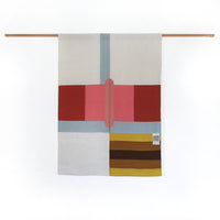 A contemporary-style Japanese noren curtain displayed on a simple wooden pole. The curtain is made of two panels featuring an abstract design of large pink, red and pale blue rectangles and stripes in the top two thirds , and a square of yellow and brown stripes in the bottom right. At the enter is a slim pink oval. Designed and made by Amy Croft, Modja Modja House and on display at NiMi Projets UK.  Image ©Amy Croft.