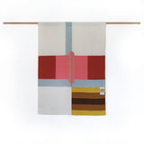 A contemporary-style Japanese noren curtain displayed on a simple wooden pole. The curtain is made of two panels featuring an abstract design of large pink, red and pale blue rectangles and stripes in the top two thirds , and a square of yellow and brown stripes in the bottom right. At the enter is a slim pink oval. Designed and made by Amy Croft, Modja Modja House and on display at NiMi Projets UK.  Image ©Amy Croft.