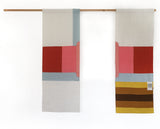 A contemporary-style Japanese noren curtain, split into two separate rectangular panels that are displayed on a wooden pole. The left panel features a white background with central red and pink rectangles above a pale blue horizontal stripe, and a pale vertical stripe in the top right. The right panel has matching pink, red and blue rectangles, with yellow and brown stripes below it.  Designed and hand made by Amy Croft, Modja Modja House, and available at NiMi Projects UK. Image ©Amy Croft.