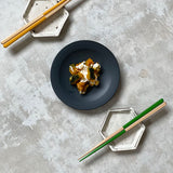 A black side plate with stewed pumpkin, served with two sets of Style of Japan, bicolour Oedo hashi chopsticks — one pair is yellow and wood, the other green and wood.