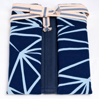 A folded contemporary-style short maekake Japanese apron in blue, featuring side panels with a white "crystal" pattern of lines, and a grey and white waist belt. On show at NiMi Projects UK.