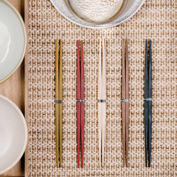 Five pairs of Style of Japan's Hashi double-end tapered wooden Ryokuchi chopsticks, lined up together on a mat with bowls in a traditional setting. Each pair is a different colour from left to right: green, red, natural, brown and black. Made in Japan and available at NiMi Projects UK.