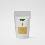 A white pouch of Awaido Japanese green tea leaves. The pouch has a rectangle label with a green illustration of a tea leaf and the name of the tea in Japanese kanji. Below the kanji is an orange section of the label with the name in English: Kuan. Image ©AWAIDO
