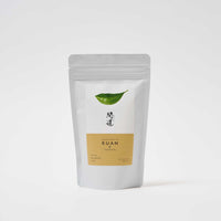A white pouch of Awaido Japanese green tea leaves. The pouch has a rectangle label with a green illustration of a tea leaf and the name of the tea in Japanese kanji. Below the kanji is an orange section of the label with the name in English: Kuan. Image ©AWAIDO