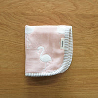 A folded, pink Fuwara multipurpose small muslin on top of a wooden surface. The muslin is made of Japanese cotton gauze and features a stitched pattern of white flamingos. plus a trimming of grey stripes.