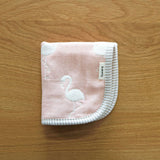 A folded, pink Fuwara multipurpose small muslin on top of a wooden surface. The muslin is made of Japanese cotton gauze and features a stitched pattern of white flamingos. plus a trimming of grey stripes.