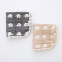 Two folded Fuwara multipurpose small muslins made of Japanese cotton gauze, one in navy blue, the other in light brown. Each one features a pattern of white woven hedgehogs and matching striped trimming.